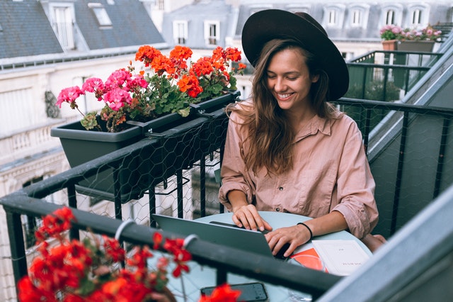 Žena v klobúku sedí na balkóne a pracuje s notebookom